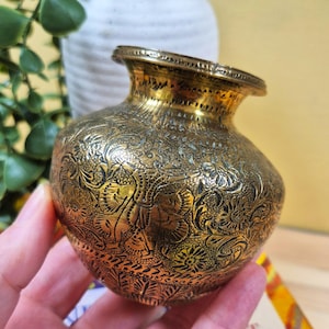 May include: A small, antique brass vase with detailed floral and leaf patterns etched across its surface. The vase has a rounded body, a flared rim, and a golden hue. The intricate design adds a touch of elegance to this decorative piece.