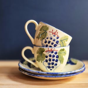 May include: Two cream-coloured teacups and saucers stacked, featuring a hand-painted grape design. The cups have curved handles and are decorated with blue grapes, green leaves, and red stems. The saucers have a matching grape design and blue trim.