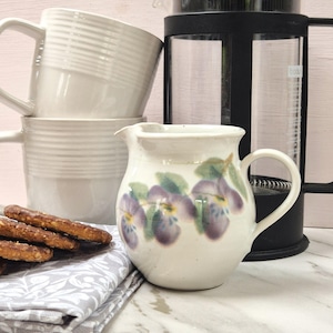 May include: A white ceramic creamer decorated with purple and yellow pansies. Two white mugs are stacked behind the creamer, and a French press is visible. Pretzels and a patterned napkin are in the foreground. A charming kitchen scene.