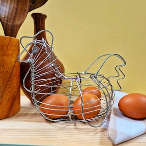 May include: A silver wire hen-shaped egg basket containing brown eggs. The basket sits on a wooden surface, with a brown vase and wooden utensils behind it. A checkered napkin is to the right.