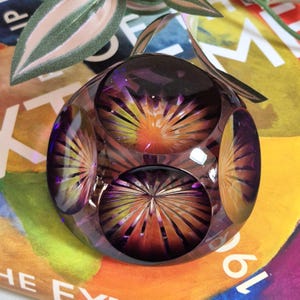 May include: A clear, round paperweight with a complex, multi-faceted design. The paperweight features a central, sunburst-like pattern in shades of orange and yellow, surrounded by a deep purple hue. The overall effect is a striking display of light and colour.