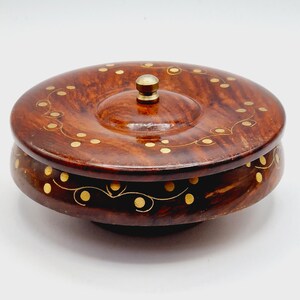 May include: A circular wooden box with a lid, featuring a dark brown finish and gold-coloured decorative accents. The lid is topped with a small, brass-coloured knob. The box is raised on a small base.