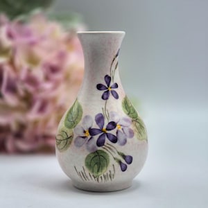 May include: A small ceramic bud vase with a pale pink base colour, featuring hand-painted purple violet flowers with yellow centres and green leaves. The vase has a narrow neck that flares slightly at the top and a rounded, bulbous body shape.