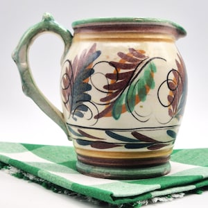 May include: A ceramic jug with a cream-coloured base, decorated with colourful leaf and swirl designs in shades of blue, orange, and green. The handle is light green, and the rim is trimmed in green. The jug sits on a green and white checkered cloth.
