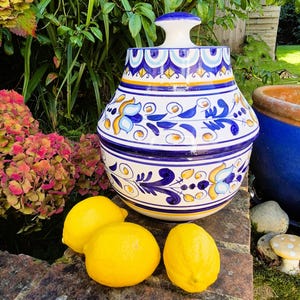 May include: A blue and white ceramic jar with a floral design and a lid. The jar is decorated with yellow and gold accents. Three yellow lemons are in front of the jar.