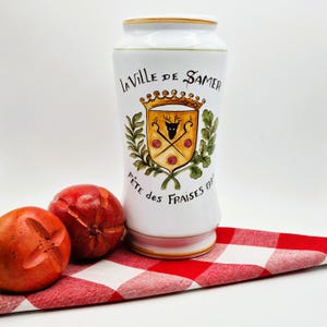 May include: A white ceramic jar with a decorative crest and the text "LA VILLE DE SAMER" and "FÊTE DES FRAISES 199". Two red fruits sit in front of the jar on a red and white checkered cloth.