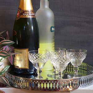 May include: A still life arrangement featuring a champagne bottle, a bottle of liqueur, and four crystal glasses on a silver tray. The glasses have a diamond pattern. A plant is visible on the left side of the image.