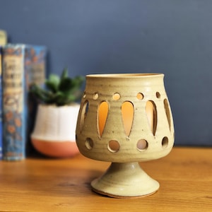 May include: A beige ceramic candle holder with a pedestal base and a flared top. The top has teardrop and circular cutouts, allowing light to shine through. The candle holder is on a wooden surface.