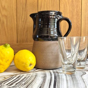 May include: A two-toned ceramic pitcher with a glossy black upper section and a matte tan base, accompanied by two clear glass tumblers and two bright yellow lemons. The pitcher features a curved handle and a spout for pouring liquids.