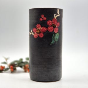 May include: A cylindrical black vase with a textured, matte finish. The front features a hand-painted design of red berries and a single green leaf, accented by small, stylised gold-coloured star shapes scattered near the botanical elements.