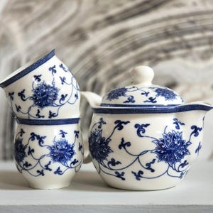 May include: A white ceramic teapot with a lid and two matching cups, all decorated with a blue floral pattern and a geometric border. The teapot features a curved handle and spout, while the two cups are stacked vertically next to the teapot.
