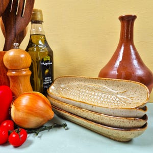 May include: Three speckled ceramic serving dishes with a textured design are stacked. A bottle of olive oil, a brown vase, and fresh vegetables, including tomatoes and an onion, are also visible.