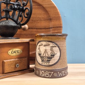 May include: A brown ceramic mug featuring a relief of a sailing ship within a circular frame, with the text 1987 WEYMOUTH inscribed at the base. Beside it stands a wooden coffee grinder with a black metal crank and a brass CAFE plaque.