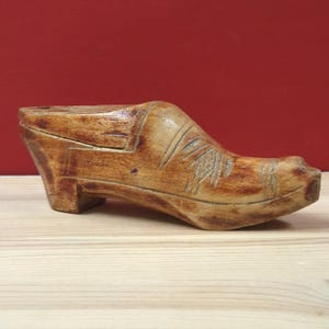 May include: A carved wooden shoe form with a light brown finish and visible wood grain. The object features a block heel, a rounded toe, and decorative etched lines across the vamp. The surface shows natural variations in colour and texture.