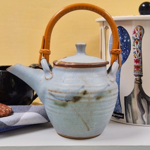 May include: A pale blue ceramic teapot with a brown leather handle and lid. The teapot has a rounded body and a spout. The handle is attached with leather straps. The teapot is on a white surface.