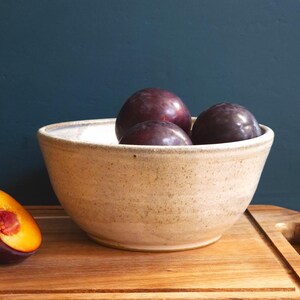 May include: A beige ceramic bowl containing three dark purple plums. A halved plum with a yellow interior sits on a wooden chopping board. The bowl has a speckled texture and a light-coloured rim. The background is a dark teal colour.
