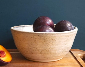 May include: A beige ceramic bowl filled with three dark purple plums. A cut plum with a yellow interior is on a wooden cutting board. The bowl has a speckled texture and a light-colored rim. The background is a dark teal color.