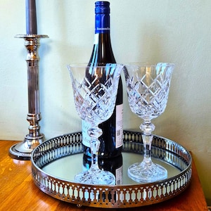 May include: A wine presentation set with a bottle of wine, two crystal wine glasses, and a mirrored tray. The wine glasses have a diamond pattern. A silver candlestick is in the background. The wine bottle has a blue cap.