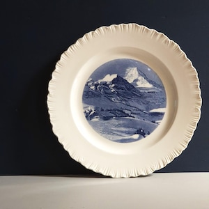 May include: A round, cream-coloured ceramic plate featuring a scalloped, textured rim. The centre of the plate displays a detailed blue and white transfer print illustration of a snow-covered mountain landscape with a small cabin in the foreground.
