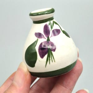 May include: A small, cream-coloured ceramic vase with a tapered neck and a rounded body. The vase features hand-painted purple floral motifs with green leaves and stems, accented by a solid dark green band around the rim and the base.