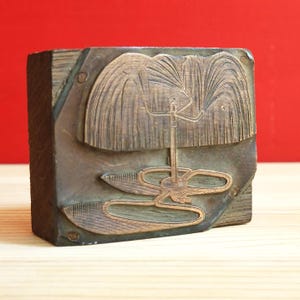 May include: A vintage wooden block with a copper relief design. The design depicts a stylized fountain with cascading water and a central figure. The block is rectangular and probably used for printing.