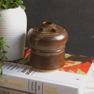 May include: A brown ceramic flower frog with a rounded top and a cylindrical base. The top has several holes for arranging flowers. The frog is on a stack of books next to a white vase with a plant.