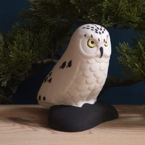 May include: A white owl figurine with black spots on its wings and head, featuring bright yellow eyes and a black beak. The owl is perched on a solid black base, showcasing a textured feather pattern across its body and a focused, forward-facing gaze.