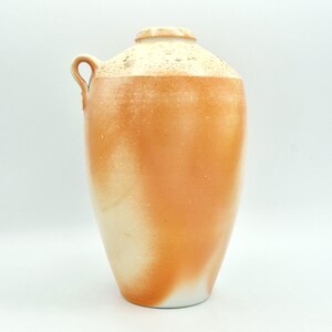 May include: A large ceramic vase with a handle, showcasing a colour gradient. The top is a speckled cream colour, blending into a warm orange, and fading to white at the base. The vase has a rounded top and a single handle.