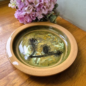 May include: A ceramic bowl with a landscape design. The bowl has a light brown rim and a green interior with a swirling pattern of yellow and white. The landscape features black trees and a body of water.