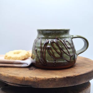 May include: A ceramic mug with a wide base and a glossy olive green glaze featuring dark brown, raised, swirling decorative lines. The mug has a sturdy handle on the right side and a smooth, flared rim. A light-coloured, round biscuit sits nearby.