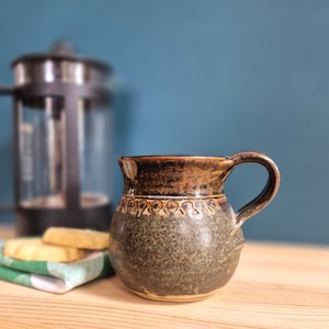 May include: A round ceramic mug with a dark green speckled base and a smooth brown glazed rim. The mug features a decorative band of small circular indentations around the upper section and a sturdy curved handle on the right side.