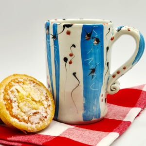 May include: A ceramic mug with a white base, blue vertical stripes, and a decorative handle. The mug features black and red abstract designs. A pastry sits on a red and white checkered cloth.