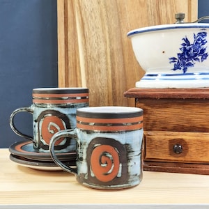 May include: Two ceramic mugs with matching saucers featuring a light blue and dark brown glaze with a prominent orange spiral design on the front. The mugs have a dark brown rim and a horizontal orange band near the top of the cylindrical body.