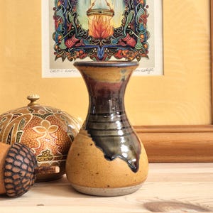 May include: A handcrafted ceramic vase with a flared top and a rounded body. The vase has a colour palette of tan, brown, and dark green, with a glossy finish. The vase is displayed with other decorative items, including a decorative acorn and a patterned ceramic container.