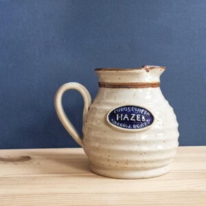 May include: A cream-coloured ceramic pitcher with a speckled finish and horizontal ribbed texture. It features a brown rim and a blue oval plaque on the front with the white text MIDDLEWICH HAZEL NARROW BOATS. The pitcher has a single handle.