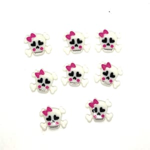 May include: A collection of eight white skull-shaped embellishments. Each skull has black heart-shaped eyes, a pink bow, and a pink mouth. The skulls are arranged on a white background.