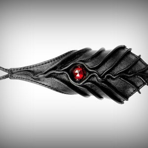Pode incluir: Pulseira de couro preto com um detalhe de pedra preciosa vermelha. A pulseira tem um design texturizado e orgânico.