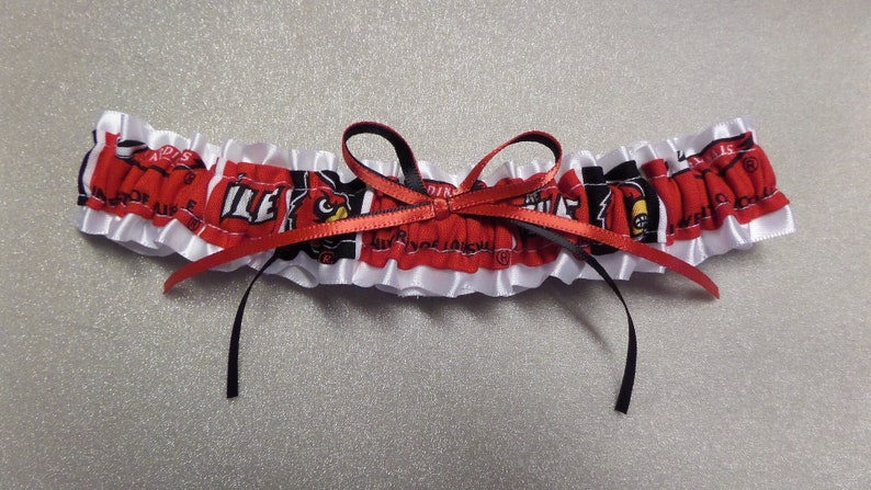 May include: White satin garter with a red and black ribbon bow. The garter features a red and black fabric with a repeated pattern of a cardinal bird and the word "Louisville".