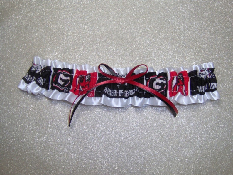 May include: White satin garter with a red and black ribbon bow. The garter features a repeating pattern of the University of Georgia logo and mascot.