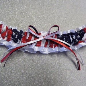 May include: A white satin garter with a red, white, and blue patterned fabric insert. The garter has a bow made of red, white, and black ribbon.