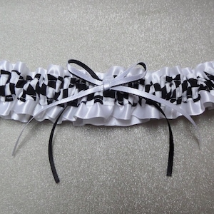May include: A garter featuring a black and white checkered pattern with a ruffled white satin trim. The center is adorned with a decorative bow made of one thin black ribbon and one thin white ribbon, with long ribbon tails hanging downward.