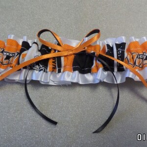 May include: A white satin garter with a black and orange fabric insert featuring a repeated pattern of a sports team logo. The garter has a black satin bow.