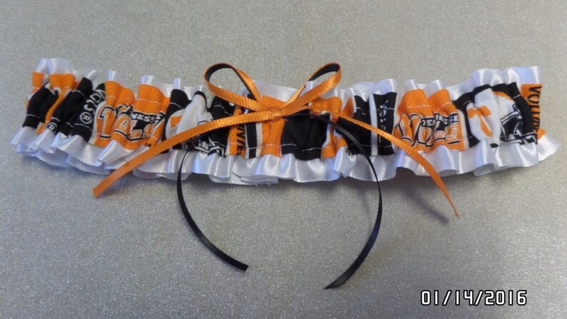 May include: A white satin garter with a black and orange fabric insert featuring a repeated pattern of a sports team logo. The garter has a black satin bow.