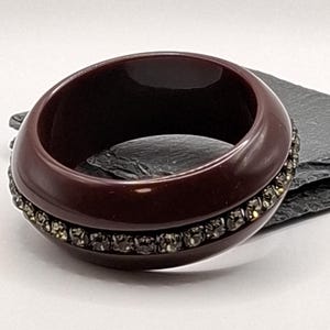 Op de afbeelding: Een brede, donker bordeauxrode armband. De armband heeft een band van kleine, sprankelende, zilverkleurige stenen. De armband heeft een solide, glad en gepolijst ontwerp.