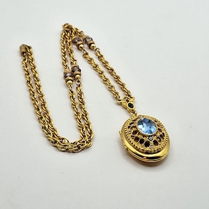 Pode incluir: Um colar com relicário em metal dourado com uma corrente torcida e pequenos detalhes de contas roxas. O relicário oval tem um design intrincado de filigrana na frente, centrado com uma pedra preciosa oval facetada em azul claro.