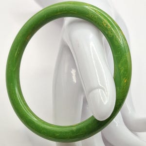 Può includere: Un braccialetto verde. Il braccialetto rotondo è di un colore verde vibrante e uniforme con sottili marmorizzazioni e una finitura liscia. Il braccialetto è esposto su una mano di manichino bianca.