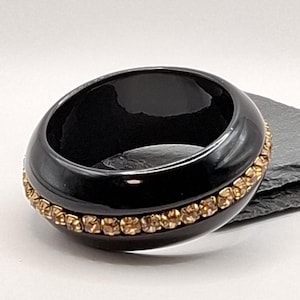 May include: A wide, black bangle bracelet with a glossy finish, adorned with a row of small, amber-toned rhinestones. The bracelet is set against a neutral backdrop.