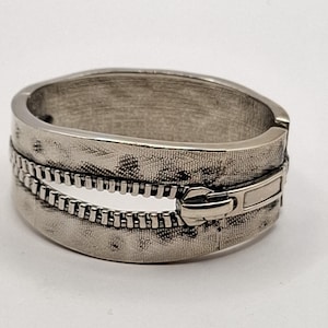 Könnte beinhalten: Ein aufklappbarer Armreif aus silberfarbenem Metall mit einer strukturierten Oberfläche, die Stoff imitiert. Das Design zeigt ein markantes Reißverschlussmotiv, das horizontal über die Mitte verläuft, inklusive eines detaillierten Reißverschluss-Schiebers.