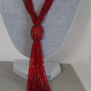 May include: A red beaded necklace with a tassel design. The necklace features multiple strands of small red beads that converge into a central, rounded bead cluster. The tassel is made of long, flowing red beads.