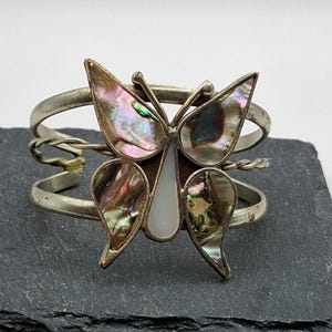 Könnte beinhalten: Ein silberfarbenes Armband mit einem zentralen Schmetterlingsmotiv. Die Schmetterlingsflügel sind mit schillernden Abalone-Muschelsegmenten in Rosa-, Grün- und Brauntönen eingelegt, während der Körper in der Mitte aus einer glatten, weißen Einlage besteht.